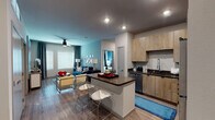 This well-appointed single-floor residence offers a thoughtfully designed open-concept layout that seamlessly connects the kitchen, dining, and living areas. The kitchen is a standout feature, showcasing light wood-grain cabinetry extending to the ceiling, dark countertops, a full suite of stainless steel appliances including a refrigerator, range, and microwave, and a central island with bar seating — ideal for casual dining and entertaining. Wood-look flooring flows throughout the common areas, while recessed lighting and a ceiling fan contribute to a bright, comfortable ambiance. Large windows allow generous natural light into the living room, which is anchored by a teal accent wall and access to the entry foyer. The primary bedroom offers plush carpeting, a ceiling fan, and a large window, leading into a well-designed en-suite bathroom with dual vanities, wood-tone cabinetry, dark countertops, and direct access to a generously sized walk-in closet with multi-tier shelving and hanging rods. A second bedroom features its own bold blue accent wall and ample natural light. The second bathroom includes a tub-shower combination with wood-look flooring, completing this stylish and functional residence.