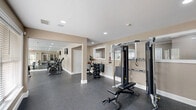 Located at 100 Cobblestone Drive in Coatesville, PA, this fitness center offers a well-appointed exercise room designed to support a comprehensive workout experience. The space features durable speckled rubber flooring throughout, providing both safety and functionality for a wide range of activities. Recessed lighting illuminates the room evenly, complemented by large windows fitted with horizontal blinds that invite natural light while offering views of the surrounding greenery outdoors.

The exercise room is thoughtfully organized into distinct zones. A dedicated cardio area houses treadmills, upright and recumbent stationary bikes, and an elliptical machine, with wall-mounted flat-screen televisions providing entertainment during workouts. A full-length mirrored wall expands the sense of space and assists with form and technique. The strength training section is equipped with a cable crossover machine, an adjustable weight bench, a dumbbell rack with a full set of free weights, and medicine balls. Structural columns with clean white bases add architectural character while defining the open layout. Neutral warm-toned walls and white trim throughout lend the space a clean, modern aesthetic. The room also includes direct exterior access through a glass-paneled door, ensuring convenient entry and exit.