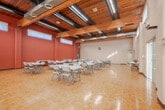 Located at 3 Leaside Drive in St. Catharines, Ontario, this single-floor event space offers a generously sized, open-concept venue well-suited for a wide range of gatherings and functions. The main hall is defined by its striking vaulted ceiling with exposed dark wood beams and warm tongue-and-groove wood paneling, lending the space a welcoming, character-filled atmosphere. Classic parquet hardwood flooring spans the entire area, adding a timeless aesthetic underfoot. Two contrasting wall finishes — painted white concrete block and terracotta-toned brick — create a distinctive, dynamic interior. Clerestory windows and tall vertical windows along the walls allow natural light to filter generously throughout the hall. The space is equipped with a projection screen, audio speakers on stands, and track lighting, making it practical for presentations, performances, and community events. Folding tables and chairs are available throughout, allowing flexible seating arrangements to suit various event formats. Decorative indoor plants are placed around the hall, softening the industrial character of the space. A pass-through service window and a service table area are accessible along one wall, providing convenient catering support. A secondary area connects to the main hall, offering additional functional space. Overall, this venue combines practical amenities with architectural warmth.