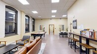 Located at 301 W 1st St in downtown Duluth, MN, this amenity space combines a vibrant social lounge with a comprehensive fitness center across a single, generously proportioned floor. The main lounge area is bright and welcoming, featuring warm yellow walls, light wood-style flooring, and a drop ceiling with recessed panel lighting. Large arched windows framed in dark wood trim—characteristic of the building's historic architecture—allow natural light to fill the space and lend a distinctive character. The lounge is furnished for both recreation and relaxation, with a full-size billiards table as a centerpiece, comfortable leather seating, and a wall-mounted television. A high-top bar counter with stools and a long conference-style table with folding chairs provide additional gathering options. The fitness center is accessible directly from the lounge and is divided into distinct zones. One area houses cardio equipment, including treadmills and an elliptical machine, alongside an exposed brick accent wall that reinforces the building's historic charm. A separate strength training area offers a multi-station cable machine, a full dumbbell rack, and adjustable benches. Mirrored wall panels and a ballet-style barre complete the workout space, catering to a range of fitness activities. Together, these spaces provide a versatile, well-appointed environment for both leisure and wellness.