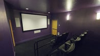 This private home theater is purpose-built to deliver a true cinema experience. The room is finished in a rich, deep purple on all walls and ceiling, a deliberate design choice that minimizes light reflection and enhances on-screen contrast for optimal viewing. A large, centrally mounted projection screen dominates the front wall, framed cleanly to serve as the room's focal point.

Seating is arranged in a tiered, stadium-style layout, ensuring clear sightlines from every position. The plush, reclining chairs are equipped with built-in cup holders, adding a practical and comfortable touch for extended viewing sessions. A metal safety railing separates the seating tiers, reinforcing both the cinematic aesthetic and functional safety of the raised rows.

Recessed ceiling lighting provides subtle ambient illumination when needed, while a wall-mounted sconce near the entry door offers a warm accent glow. A ventilation unit is visible along the lower wall, ensuring the space remains comfortable during use. The natural wood-toned door provides a warm contrast against the dark interior palette. Dark carpeting throughout the room further absorbs sound and light, contributing to the theater's acoustically considerate and visually immersive design.