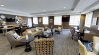 Located at 225 Echelon Rd in Voorhees, NJ, this single-floor community space is designed with both comfort and functionality in mind. The main lounge area serves as the centerpiece of the space, offering a well-appointed gathering environment with a thoughtfully curated mix of seating arrangements. Plush sofas and accent chairs — including boldly patterned upholstered pieces — are arranged around dark wood coffee and side tables, creating inviting conversation areas suited for both casual relaxation and social gatherings. The room features a tray ceiling with recessed lighting, lending a refined architectural detail that adds depth and visual interest overhead. Rich, dark brown walls provide a warm, sophisticated backdrop, complemented by patterned carpet flooring that adds texture underfoot. A wall-mounted flat-screen television anchors one end of the room, while roller shades on multiple windows allow for flexible control of natural light. A dining area is visible toward the rear, extending the functionality of the space for multiple uses. The overall ambiance is modern yet inviting, balancing neutral tones with bold accent pieces to create a polished, welcoming environment suitable for a variety of activities.