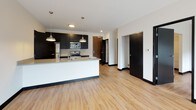 Unit 221 is a thoughtfully designed single-floor residence that blends contemporary aesthetics with practical functionality throughout. Light wood-tone flooring flows consistently across all rooms, complemented by crisp white walls and bold black trim details that create a clean, modern contrast.

The open-concept kitchen serves as a central hub, outfitted with dark cabinetry, stainless steel appliances including a refrigerator, range, and microwave, and a white subway tile backsplash. A spacious island with a built-in sink and pendant lighting overhead offers both additional prep space and casual seating.

The living area is generously proportioned, featuring large black-framed windows and a full sliding glass door that opens onto a private balcony with outdoor views, allowing abundant natural light to fill the space. A secondary bedroom includes two large windows and direct access to a walk-in closet equipped with built-in shelving and hanging rods.

The bathroom showcases a sleek, modern design with floor-to-ceiling dark gray tile in the walk-in shower, a rain showerhead, a gray vanity with ample storage, and wood-tone flooring. An in-unit stacked washer and dryer provide added convenience. The overall ambiance is contemporary and cohesive throughout.