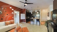 This single-floor space showcases a thoughtfully designed open-concept layout that integrates the living, dining, and kitchen areas into one cohesive environment. The living area is anchored by plush neutral carpeting and features a striking deep-red accent wall adorned with flowing abstract line artwork, adding a bold artistic focal point to the room. A sleek ceiling fan with a central light fixture overhead provides both airflow and ambient lighting.

The kitchen is outfitted with dark cabinetry, stainless steel appliances including a refrigerator and microwave, and light-toned countertops. A peninsula-style breakfast bar with tall dark bar stools creates a natural transition between the kitchen and the living space, making it ideal for casual dining or entertaining. Pendant lighting hangs above the bar, complementing the track lighting installed in the kitchen for functional task illumination.

The overall ambiance balances contemporary design with warm, inviting tones. The contrast between the dark kitchen cabinetry and the warm red accent wall creates visual interest, while the open layout ensures the space feels connected and welcoming throughout.