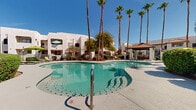 Located at 1891 N Litchfield Rd in Goodyear, Arizona, this outdoor amenity area showcases a generously sized freeform swimming pool as its centerpiece. The pool's curvilinear design adds a resort-style character to the space, with crystal-clear turquoise water reflecting the abundant Arizona sunshine. The surrounding pool deck is finished in a textured aggregate concrete surface, providing both durability and a non-slip walking area for residents and guests.

Tall, mature palm trees frame the space, lending a tropical ambiance while offering natural shade throughout the day. Lush desert-adapted landscaping, including neatly trimmed hedges, flowering shrubs, and gravel ground cover, lines the perimeter and contributes to a well-maintained, inviting atmosphere.

Multiple lime-green lounge chairs are positioned along the deck, offering comfortable spots for sunbathing and relaxation. A covered cabana structure with a tiled roof provides a shaded retreat, ideal for escaping the desert heat. Wrought-iron fencing encloses the pool area, ensuring safety and privacy.

The surrounding multi-story buildings feature Southwestern-style stucco architecture with neutral tones that complement the natural desert palette. The open-air layout, abundance of natural light, and carefully curated landscaping combine to create a welcoming and functional outdoor recreational space.
