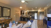 This single-floor unit offers an open-concept layout that connects the living, dining, and kitchen areas into one cohesive, functional space. The living area is anchored by a comfortable seating arrangement beneath a ceiling fan with integrated lighting, contributing to both comfort and air circulation. Hardwood-style flooring runs throughout, adding warmth and visual continuity across the space.

The kitchen is a standout feature, outfitted with dark cabinetry, stainless steel appliances including a refrigerator, range, and microwave, and a mosaic tile backsplash that adds a decorative accent. A peninsula with a granite-style countertop provides additional prep space and casual seating with industrial-style bar stools, making it ideal for entertaining or everyday dining.

Recessed lighting and pendant fixtures illuminate the kitchen, while the open sightlines allow natural light to travel freely through the space. Neutral wall tones complement the darker cabinetry and blue accent décor. Multiple doorways are visible, suggesting access to additional rooms beyond the main living area. Overall, the space balances modern aesthetics with practical, everyday functionality.