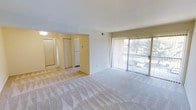 This single-floor residence offers a clean, functional layout with a spacious open-plan living area at its heart. The living room is finished with plush wall-to-wall carpeting in a neutral tone, creating a comfortable and cohesive feel throughout. A large sliding glass door fitted with vertical blinds dominates one wall, allowing generous natural light to fill the space while providing direct views of the surrounding outdoor greenery and parking area beyond.

The living area flows openly into an adjoining space that houses a white refrigerator, suggesting a kitchen or dining transition zone. Warm yellow-toned walls in this connected area provide a subtle contrast to the bright white of the main living space. Multiple bi-fold and panel doors are visible, indicating ample closet and storage options nearby.

Overhead, a flush ceiling light fixture adds functional illumination to the secondary area, while the main living room benefits primarily from the abundant natural light provided by the sliding glass door. The overall ambiance is bright, clean, and move-in ready, with a straightforward layout suited for comfortable everyday living.