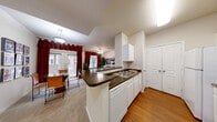 This single-floor residence offers a thoughtfully designed open-concept layout that seamlessly connects the kitchen, dining, and living areas. The kitchen is well-equipped with white cabinetry, dark countertops, a double-basin sink, a full suite of white appliances including a range, refrigerator, and dishwasher, and durable hardwood flooring that extends into the main corridor. A peninsula counter serves as a natural divider between the kitchen and the adjacent dining area, which features a glass-top dining table and access to the outdoors through French doors dressed in rich red curtains. The living room is a cozy, carpeted space with a ceiling fan, large windows offering natural light, and built-in display shelving. The bedrooms are carpeted and tranquil, each featuring ceiling fans, ample natural light, and vaulted ceiling accents in select areas. The bathroom is clean and functional, appointed with tile flooring, a single-vanity sink with white cabinetry, and a tub-shower combination with green tile accent detailing. Throughout the residence, a neutral warm color palette, consistent trim work, and ceiling fans in key rooms contribute to a comfortable and cohesive living environment.