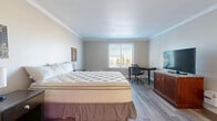 Located at 2886 S Circle Dr in Colorado Springs, CO, this efficiently designed studio unit offers a well-organized single-floor layout that maximizes every square foot. The main living area serves as both bedroom and living space, featuring light gray wood-look laminate flooring, neutral-toned walls, and elegant crown molding that adds a refined architectural touch throughout. Natural light filters in through a window fitted with blinds, brightening the open room. The space accommodates a king-sized bed, two nightstands, a dedicated work desk with an ergonomic chair, and a media console — providing a comfortable and functional environment. A compact kitchenette is tucked into one corner of the main room, outfitted with white cabinetry, a microwave, a sink, and a tiled backsplash, offering convenient in-unit food preparation. The bathroom features light ceramic tile flooring, a vanity with a granite-style countertop, and a full bathtub and shower combination complete with a safety grab bar and a curtained enclosure. The entrance flows naturally into the main living area, contributing to the unit's cohesive and practical design. Overall, the space presents a clean, neutral aesthetic suited for comfortable single-occupancy living in Colorado Springs.