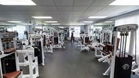 This single-floor fitness facility offers a generously sized, open workout space designed to accommodate a wide range of strength training activities. The room is outfitted with an extensive collection of Atlantis-brand resistance machines, systematically arranged in rows to maximize usability and traffic flow throughout the floor. Equipment includes cable machines, leg press stations, chest and back machines, and a variety of other targeted strength training apparatus, providing users with a thorough full-body workout experience. The flooring consists of dark rubber matting, a practical and durable surface well-suited for heavy equipment and high-traffic use. Overhead, a suspended drop ceiling hosts rows of recessed fluorescent panel lighting, ensuring consistent, even illumination across the entire space. Full-length mirrors line the walls, visually expanding the room while allowing users to monitor their form during exercise. The overall ambiance is clean, functional, and professional, reflecting a facility focused on delivering a structured and efficient training environment.