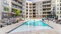 Located in the vibrant Dilworth neighborhood of Charlotte, NC, this modern multi-story property at 536 W Tremont Ave showcases a thoughtfully designed courtyard amenity space that serves as a central gathering hub. The architecture features a contemporary exterior with neutral grey siding, dark brick accents, and large windows, with private balconies lining multiple floors and overlooking the courtyard below.

At the heart of the outdoor space is a generously sized resort-style swimming pool with crystal-clear blue water, complemented by a separate spa. The expansive pool deck is finished with large-format natural stone pavers, providing an upscale and durable surface throughout. Surrounding the pool, a well-appointed collection of modern sling lounge chairs with coordinating side tables offers ample space for relaxation and sunbathing. Patio umbrellas are positioned throughout the deck for shade, while strategically placed lamp posts add ambient lighting for evening use.

Lush landscaping with young trees and greenery is integrated into the courtyard design, softening the hardscape and enhancing the outdoor atmosphere. A dedicated lounge seating area with sofas and chairs is tucked along the building's perimeter, creating a comfortable social zone separate from the pool deck. Ground-floor access points connect the courtyard to interior common areas, ensuring seamless indoor-outdoor flow throughout this well-appointed property.