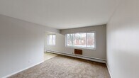 Located at 2928 Kenwick Cir in Lansing, Michigan, this single-floor, 2-bedroom, 1-bathroom home offers a practical and straightforward layout. The living room is a well-proportioned space featuring neutral-toned carpet, clean white walls, and multiple windows that allow natural light to fill the room. Baseboard heating runs along the exterior walls, and a wall-mounted AC unit provides additional climate control. The kitchen features warm wood cabinetry, tile flooring, and a window overlooking the outdoors, creating a functional cooking space. A hallway connects the main living areas to the bedrooms, with built-in shelving visible at the end. The primary bedroom is a generously sized carpeted room with neutral gray walls and a door leading to a dedicated closet with shelving. The second bedroom also features carpet, a window, and a built-in closet with louvered bifold doors for ample storage. The bathroom is finished with classic white square tile on the walls accented by subtle green tile details, a mosaic tile floor, a tub-shower combination, and a wood-trimmed vanity with a mirror. Throughout the home, a consistent neutral color palette and carpeted flooring create a cohesive and functional living environment.