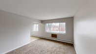Located at 2928 Kenwick Cir in Lansing, Michigan, this single-floor, 2-bedroom, 1-bathroom home offers a practical and straightforward layout. The living room is a well-proportioned space featuring neutral-toned carpet, clean white walls, and multiple windows that allow natural light to fill the room. Baseboard heating runs along the exterior walls, and a wall-mounted AC unit provides additional climate control. The kitchen features warm wood cabinetry, tile flooring, and a window overlooking the outdoors, creating a functional cooking space. A hallway connects the main living areas to the bedrooms, with built-in shelving visible at the end. The primary bedroom is a generously sized carpeted room with neutral gray walls and a door leading to a dedicated closet with shelving. The second bedroom also features carpet, a window, and a built-in closet with louvered bifold doors for ample storage. The bathroom is finished with classic white square tile on the walls accented by subtle green tile details, a mosaic tile floor, a tub-shower combination, and a wood-trimmed vanity with a mirror. Throughout the home, a consistent neutral color palette and carpeted flooring create a cohesive and functional living environment.