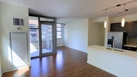 This single-floor unit presents a clean, contemporary layout centered around an open living area with rich hardwood flooring that runs throughout. Large floor-to-ceiling sliding glass doors flood the space with natural light while providing direct access to a private balcony, offering views of the surrounding urban streetscape and neighboring brick buildings. The living area is generously proportioned, with white walls and a neutral color palette that enhance the sense of openness and light.

Adjacent to the living space, a partial-height dividing wall defines the kitchen area without fully enclosing it, preserving the open-concept flow. Pendant lighting hangs above what appears to be a kitchen counter or breakfast bar, adding a modern touch to the layout. The kitchen area features a pass-through design that connects it visually to the main living space.

The overall ambiance is modern and urban, with minimal ornamentation allowing the architectural details — including the clean lines of the window framing, the sliding glass door system, and the polished flooring — to take center stage. The space is well-suited for those seeking a bright, functional, and contemporary urban living environment.