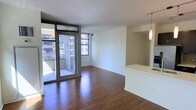 This single-floor unit presents a clean, contemporary layout centered around an open living area with rich hardwood flooring that runs throughout. Large floor-to-ceiling sliding glass doors flood the space with natural light while providing direct access to a private balcony, offering views of the surrounding urban streetscape and neighboring brick buildings. The living area is generously proportioned, with white walls and a neutral color palette that enhance the sense of openness and light.

Adjacent to the living space, a partial-height dividing wall defines the kitchen area without fully enclosing it, preserving the open-concept flow. Pendant lighting hangs above what appears to be a kitchen counter or breakfast bar, adding a modern touch to the layout. The kitchen area features a pass-through design that connects it visually to the main living space.

The overall ambiance is modern and urban, with minimal ornamentation allowing the architectural details — including the clean lines of the window framing, the sliding glass door system, and the polished flooring — to take center stage. The space is well-suited for those seeking a bright, functional, and contemporary urban living environment.