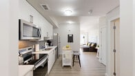Located at 7711 MacLeod Trail SW in Calgary, this thoughtfully designed one-bedroom, one-bathroom unit makes efficient use of its compact footprint. The open kitchen anchors the living space with crisp white cabinetry, a grey subway tile backsplash, stainless steel appliances including a range, microwave, and dishwasher, and light wood-look flooring that carries throughout. A portable butcher block island with stools adds a practical casual dining option, maximizing versatility in the layout. The bedroom is a standout, featuring a tray ceiling with recessed lighting that lends a sense of height and refinement. Framed artwork adorns the walls, and French patio doors open directly onto a private outdoor space, flooding the room with natural light and offering pleasant green views of the landscaped exterior grounds. The bathroom is clean and functional, finished with a white vanity, wood-look tile flooring, and a full tub-shower combination with grey tile surround. The building exterior showcases a contemporary multi-storey design with stone and glass detailing, complemented by well-maintained garden beds, glass-railed walkways, and open-air common areas. This unit offers a well-appointed, modern living experience in a vibrant Calgary location.