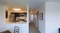 This single-floor residence offers a clean, contemporary layout designed for efficient living. The kitchen is a standout feature, equipped with dark espresso-toned cabinetry that contrasts sharply against light countertops and a white subway tile backsplash. Stainless steel appliances, including a refrigerator, microwave, and gas range, provide a sleek, functional workspace. A breakfast bar with seating extends from the kitchen, creating a casual dining area that also serves as a natural divider between the kitchen and the adjoining living space.

A carpeted hallway extends from the main living area, lined with multiple doors leading to additional rooms. The corridor is well-lit by a flush-mount ceiling fixture and is flanked by neutral white walls, giving the space a bright and airy feel. A framed piece of wall art adds a subtle decorative touch along the hallway. At the far end, a glimpse into one of the rooms reveals natural light, suggesting a bright interior. The overall design emphasizes a neutral color palette, modern finishes, and a practical flow throughout the home.