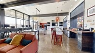 This vibrant, single-floor community clubhouse is thoughtfully designed to accommodate a variety of social and productive activities within a generously sized open-plan layout. Gleaming polished terrazzo floors stretch throughout, reflecting abundant natural light that pours in through expansive floor-to-ceiling windows and glass doors, offering views of the surrounding outdoor areas, including a pool.

The space is divided into several well-defined zones. A comfortable lounge area features plush sofas and armchairs arranged around a round coffee table, anchored by a branded wall medallion that adds a bold focal point. Adjacent to this, a games area includes a full-sized pool table and classic arcade machines, creating an energetic entertainment hub.

A dedicated co-working station lines one wall with desktop computers and bar-height seating, providing a functional workspace flooded with natural light. A central high-top dining table with vibrant red bar stools serves as a social gathering point beneath a striking pendant light fixture. Built-in shelving and wall-mounted flat-screen TVs enhance the entertainment options throughout.

Enclosed glass-walled rooms labeled "Study" and "Play" offer semi-private spaces for focused work or recreation. Exposed structural beams and a warm, contemporary color palette of deep reds, charcoals, and neutrals give the space a modern, lively character.