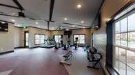 This fitness facility is thoughtfully designed to support a wide range of workout routines across two distinct areas. The main fitness floor features soaring ceilings accented by dark exposed beam detailing, recessed lighting, and a ceiling fan that contribute to a spacious, energizing atmosphere. Large windows along multiple walls invite abundant natural light while offering outdoor views, keeping the environment bright and open. The flooring incorporates a bold burgundy and light gray patterned carpet layout that defines different workout zones throughout the room. Equipment includes treadmills, elliptical trainers, a rowing machine, a rack of free weights, and strength training machines. Wall-mounted flat-screen televisions provide entertainment throughout the workout. Large mirrors along one wall expand the sense of space and allow users to monitor their form during strength exercises. The dedicated spin and yoga room offers a more intimate setting, featuring dark hardwood-style flooring, exposed dark beam ceilings, and a dramatic full-wall mural of a mountain cyclist that energizes the space. Spin bikes are arranged for group or solo sessions, accompanied by a wall-mounted screen and a window that provides natural light. The two spaces together create a comprehensive, well-appointed fitness environment suited for cardio, strength training, cycling, and yoga.