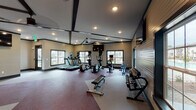 This fitness facility is thoughtfully designed to support a wide range of workout routines across two distinct areas. The main fitness floor features soaring ceilings accented by dark exposed beam detailing, recessed lighting, and a ceiling fan that contribute to a spacious, energizing atmosphere. Large windows along multiple walls invite abundant natural light while offering outdoor views, keeping the environment bright and open. The flooring incorporates a bold burgundy and light gray patterned carpet layout that defines different workout zones throughout the room. Equipment includes treadmills, elliptical trainers, a rowing machine, a rack of free weights, and strength training machines. Wall-mounted flat-screen televisions provide entertainment throughout the workout. Large mirrors along one wall expand the sense of space and allow users to monitor their form during strength exercises. The dedicated spin and yoga room offers a more intimate setting, featuring dark hardwood-style flooring, exposed dark beam ceilings, and a dramatic full-wall mural of a mountain cyclist that energizes the space. Spin bikes are arranged for group or solo sessions, accompanied by a wall-mounted screen and a window that provides natural light. The two spaces together create a comprehensive, well-appointed fitness environment suited for cardio, strength training, cycling, and yoga.