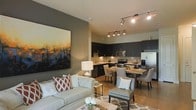This single-floor space features an open-concept layout that combines the living, dining, and kitchen areas into one cohesive, functional environment. The living area is anchored by a plush, light-toned sofa accented with patterned throw pillows and a geometric area rug, complemented by a large, vibrant abstract painting that serves as a striking focal point on the accent wall. Track lighting runs across the ceiling, providing flexible, directed illumination throughout the space. The kitchen is outfitted with dark cabinetry, stainless steel appliances, a textured stone tile backsplash, and under-cabinet lighting that highlights the countertops. A compact dining area bridges the kitchen and living space, furnished with a wooden table and upholstered chairs that maintain the space's refined aesthetic. Light hardwood-style flooring flows continuously throughout, adding warmth and visual continuity. The overall ambiance is contemporary and well-appointed, balancing dark and light tones with curated décor to create a space that feels both stylish and inviting.