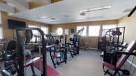 Located at 98 Oak Street in Lindenwold, New Jersey, this fitness center offers a dedicated single-floor workout space designed to support a full range of exercise routines. The room features warm, earth-toned walls that create an energizing yet welcoming atmosphere, complemented by neutral carpeted flooring throughout. Natural light streams in through multiple windows, including a distinctive circular porthole-style window, keeping the space bright and ventilated. Overhead, recessed ventilation and lighting fixtures maintain a clean, functional ceiling layout. The facility is outfitted with an impressive array of professional-grade equipment, including cable and weight machines, a multi-station strength system with adjustable benches and attachments, and cardiovascular machines such as treadmills and incline trainers. The equipment is arranged to maximize floor space while allowing comfortable movement between stations. Wall-mounted informational guides are posted to assist users with proper form and technique. This well-maintained fitness facility provides everything needed for a comprehensive workout experience in a compact yet thoughtfully organized environment.