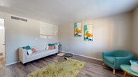 This single-floor unit offers a well-organized layout across multiple rooms, each finished with durable wood-look flooring and neutral wall tones that create a cohesive, modern atmosphere throughout.

The living room serves as the central gathering space, featuring a textured ceiling, plantation-shuttered windows that filter natural light, and an open feel well-suited for both relaxing and entertaining. Colorful abstract artwork adorns the walls, complementing the mid-century modern furnishing aesthetic. A green shag area rug anchors the seating area, which accommodates both a sofa and accent chairs.

The kitchen is efficiently designed with white cabinetry, a gray tile mosaic backsplash, granite-look countertops, and white appliances including a gas range, dishwasher, and refrigerator. A double sink and under-cabinet range hood add to the functional layout.

The bedroom is a comfortable, windowed retreat with a ceiling fan, neutral tones, and hardwood-style flooring. A secondary room functions well as a home office or flex space, also benefiting from a ceiling fan and natural light through a window with blinds.

The bathroom includes a vanity with a speckled countertop, a large mirror, and a tub-shower combination. A closet with bifold doors is visible in the floor plan overview, adding practical storage to the unit.