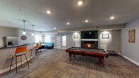 This single-floor communal space is designed to serve as a social and recreational hub, combining multiple functional zones within an open, well-lit layout. The main game room area is anchored by a full-size billiard table positioned atop patterned carpet tiles, which lend a modern, textured look to the floor. A gas fireplace set within a dark tile surround serves as a focal point on one wall, accompanied by a wall-mounted flat-screen television framed by sconce lighting and decorative artwork. Recessed ceiling lights illuminate the space evenly throughout.

Adjacent to the game room, a casual lounge area features teal upholstered seating, bar-height chairs, and pendant lighting, creating an inviting spot for informal gatherings. A cue rack and chalkboard wall accent add functional and decorative detail to the game zone.

The kitchen area is fully equipped with stainless steel appliances, including an electric range and a double-basin sink, set against white cabinetry with dark countertops. A curved countertop peninsula defines the kitchen's boundary. Two wall-mounted flat-screen televisions are positioned above the counters, and track lighting runs along the ceiling, ensuring comprehensive illumination throughout this practical yet social cooking and entertaining space.