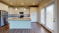 Located in Draper, Utah, this two-story home offers a thoughtfully designed layout across two levels. The main floor showcases a sweeping open-concept living and kitchen area with soaring vaulted ceilings and large windows that flood the space with natural light and provide views of the surrounding neighborhood. Rich hardwood-style flooring runs throughout the main level, complementing the clean, neutral color palette.

The kitchen is a standout feature, centered around a large island with granite countertops and an undermount sink. White shaker-style cabinetry extends to the ceiling, offering generous storage, while stainless steel appliances—including a range, microwave, and refrigerator—add a modern touch. Pendant lighting above the island and recessed ceiling lights complete the polished look. A sliding glass door connects the kitchen area to the exterior.

A convenient half bathroom on the main level features granite counters and wood-look flooring. Upstairs, carpeted bedrooms offer a comfortable retreat, with large windows providing ample natural light. The upper-level bathrooms are well-appointed, including one with a double granite vanity and another with a full tub-shower combo, both featuring white cabinetry and wood-look flooring. A wide upper hallway with clean white railings connects the upper rooms with ease.