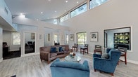 This thoughtfully designed single-story space combines open, airy architecture with functional professional interiors. The most striking feature of the main area is its dramatic vaulted ceiling, which soars to impressive heights and is lined with a continuous band of clerestory windows that flood the space with natural light and frame views of surrounding mature trees. Light-toned wood-look tile flooring runs throughout, lending a clean, cohesive aesthetic across all areas.

The main lounge area is generously proportioned, furnished with plush blue velvet seating and a curved brown sofa arranged around a glass-top coffee table, creating a welcoming and sophisticated atmosphere. Framed artwork adorns the soft gray walls, adding a curated, professional touch. A reception desk with a warm cherry wood finish anchors one side of this open space, with a wall-mounted flat-screen display and rustic wood credenza nearby.

A secondary lounge area features standard ceiling heights, cooler blue-gray walls, large picture windows overlooking greenery, and additional blue velvet seating, offering a quieter, more intimate setting. A dedicated office area includes a workstation with large windows providing outdoor views, complemented by a small seating arrangement for consultations. Throughout, recessed lighting and abundant window exposure ensure a bright, professional, and inviting ambiance.