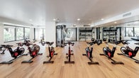This thoughtfully designed fitness facility offers a comprehensive workout environment spread across a single open floor. The space is anchored by light hardwood flooring throughout, contributing to a clean, modern aesthetic that is both functional and visually inviting.

The cardio zone is prominently positioned along a wall of large picture windows that frame lush, tropical garden views, allowing natural light to flood the area. This section houses multiple treadmills and stationary bikes, creating an energizing environment for cardiovascular training.

A dedicated cycling studio accommodates a full row of spin bikes, complemented by a large flat-screen display and floor-to-ceiling mirrors along one wall, enhancing the group fitness experience. Built-in shelving provides organized storage for accessories and equipment.

A multi-functional cable and strength training machine anchors the strength training area, alongside TRX suspension straps and a wall-mounted accessory rack stocked with kettlebells and resistance tools. Full-length mirrors throughout the space support proper form and technique.

Recessed lighting combined with abundant natural light ensures the entire facility remains bright and welcoming. The open layout allows for flexible use, accommodating individual training, group classes, and functional fitness sessions with ease. The overall ambiance is modern, clean, and professional.