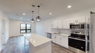 This contemporary single-floor unit showcases a clean, minimalist design with a bright, open-concept layout. The kitchen serves as a central highlight, featuring crisp white cabinetry with matte black hardware, white quartz countertops, and a chevron herringbone tile backsplash. A large central island provides additional workspace and casual dining, while stylish globe pendant lights add a modern decorative touch. Stainless steel appliances — including a refrigerator, range, microwave, and dishwasher — complete the well-equipped kitchen. Recessed lighting and large black-framed windows allow natural light to fill the space, offering views of the surrounding neighborhood. Wood-look flooring runs consistently throughout, grounding the interiors with warmth and continuity. The bedroom is a generously sized, light-filled room with a dedicated window framing urban views. A spacious walk-in closet with full-width wire shelving provides ample storage. The bathroom maintains the unit's minimalist character with white walls, a single-sink vanity with black hardware, a tub-shower combo, and matching wood-look flooring. Overall, the space presents a cohesive, modern aesthetic with thoughtful design details throughout.