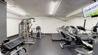 This fitness center is organized across a single floor and divided into distinct functional zones, offering a comprehensive workout environment. The cardio area is lined with a variety of machines, including treadmills, ellipticals, and stationary bikes, all positioned in front of large mirrored walls that amplify the sense of space. Ceiling-mounted flat-screen televisions are strategically placed to keep users entertained during their workouts. Bright, linear fluorescent lighting illuminates the space evenly throughout.

The strength training section features a range of selectorized cable and weight machines, a flat utility bench, and a tiered dumbbell rack stocked with multiple weight increments, supported by an open storage shelf for accessories. A dedicated free-weight and functional training area with exposed ductwork overhead gives this zone an industrial aesthetic, complemented by dark gray walls and durable rubber flooring that provides traction and impact absorption across the entire facility.

Mirrored wall panels are used extensively throughout, enhancing spatial awareness during exercise. The overall ambiance is practical and focused, designed to support a full range of fitness activities from cardiovascular conditioning to resistance and functional training.
