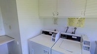 This single-floor shared laundry facility is designed with practicality and ease of use in mind. The room features a pair of coin-operated Speed Queen commercial-grade washing machines and dryers, well-known for their durability and reliable performance. Positioned side by side, the machines are accessible and straightforward to operate, making laundry tasks efficient for users.

Above the machines, white overhead storage cabinets provide additional space for supplies or personal items, keeping the area organized and clutter-free. The walls are finished in a clean, light tone that contributes to a bright and tidy atmosphere throughout the room.

Informational notices are clearly posted on the wall, offering usage guidelines and important instructions to ensure the facility is used properly and maintained in good condition. The overall layout of the space is simple and utilitarian, prioritizing functionality. The clean, white aesthetic of both the appliances and the surrounding surfaces reinforces a hygienic and well-maintained environment. This laundry facility serves as a convenient, no-frills amenity that meets everyday laundry needs effectively.
