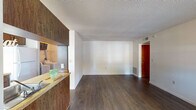 This single-floor residence offers a practical and functional layout across several well-defined living spaces. The open living area is finished with dark hardwood-style flooring that extends throughout the main areas, complemented by neutral white walls and a textured ceiling. A window allows natural light to fill the space, enhancing its clean, open feel. A half-wall partition separates the living area from the kitchen, maintaining a sense of visual connection between the two spaces.

The galley-style kitchen is efficiently designed, featuring warm wood cabinetry with ample upper and lower storage, light butcher block-style countertops, a stainless steel sink, a white electric range, and a full-size refrigerator. A window above the counter provides natural light and ventilation. The kitchen opens conveniently toward the main living space.

The bedroom is carpeted and comfortably sized, with a window offering an exterior view and allowing natural light in. A dedicated laundry area houses a full-size washer and dryer with overhead wire shelving for additional storage.

The bathroom includes a single-sink vanity with wood cabinetry, a large mirror, a full tub/shower combination with tiled surround, and hardwood-style flooring, completing a cohesive and practical interior.