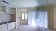 This single-floor residence offers a thoughtfully designed layout that blends comfort and functionality. The living area is bathed in natural light, thanks to large sliding glass doors dressed with vertical blinds that allow for flexible light control and outdoor access. A charming bay window adds architectural interest while further brightening the space with soft, diffused light.

Crown molding runs throughout the main living areas, lending an elegant, polished finish to the neutral-toned walls and smooth ceilings. The flooring features plush carpeting, contributing to a warm and inviting atmosphere underfoot. Decorative columns with detailed millwork serve as a subtle architectural divider, adding character and a sense of structure without closing off the open feel of the space.

Adjacent to the living area, a built-in shelving unit and countertop space — finished with a dark surface — provide practical storage and display options. The overall color palette is light and neutral, with warm beige tones on the walls complementing the soft carpeting, creating a cohesive and calming aesthetic throughout. The space is well-suited for comfortable everyday living, combining classic architectural details with practical, livable design.