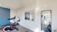 Located at 2350 SE Blackburn Rd in Cleveland, TN, this fitness room is a thoughtfully designed, single-level workout space that maximizes functionality within a compact footprint. The room features crisp white walls accented by a bold blue feature wall, creating a clean and energizing atmosphere. Durable, low-pile carpet tiles cover the floor throughout, providing a practical surface suited for a variety of exercises.

The space is well-equipped with a range of fitness equipment, including a treadmill, a Spirit-brand spin bike, a multi-position adjustable weight bench, a tiered dumbbell rack with a full set of free weights, a medicine ball rack, and a wall-mounted exercise mat. A full-length mirror mounted on one wall aids in form-checking during workouts.

Natural light enters through multiple windows, including one that overlooks a neighboring common area via an interior pass-through window, adding an open, connected feel to the room. Flush-mount circular ceiling lights and a wall-mounted split HVAC unit ensure year-round comfort. A fire extinguisher is also present, reflecting attentiveness to safety standards. Overall, this fitness room delivers a focused, practical environment for residents seeking convenient on-site exercise facilities.