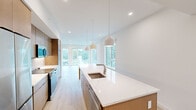 This well-designed single-floor residence in Poughkeepsie, NY offers a thoughtfully arranged layout encompassing two bedrooms, two bathrooms, a living room, kitchen, laundry room, closet, and hallway. The open-plan kitchen and living area serves as the heart of the home, highlighted by a striking elongated island topped with veined white quartz countertops and an undermount sink with a brushed nickel faucet. Pendant lighting hangs above the island, complementing recessed ceiling lights throughout. Flat-panel cabinetry in warm wood tones pairs with stainless steel appliances, including a refrigerator, range, and microwave. Light hardwood flooring extends across the main living areas, enhancing the bright, airy atmosphere. Large windows and glass doors flood the space with natural light and frame views of surrounding greenery. The primary bedroom features a well-organized closet with built-in shelving. Both bedrooms offer serene wooded views through generously sized windows. The bathrooms are finished with tile flooring and glass-enclosed walk-in showers with rainfall showerheads and floating wood shelving. A dedicated laundry room houses a full-size washer and dryer beneath a quartz shelf, adding practical convenience to this clean, contemporary residence.
