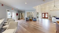 Located at 63 Mansion Blvd in Delmar, NY, this single-floor community space offers a generously sized layout with distinct areas designed for relaxation, fitness, and productivity. Throughout, light hardwood flooring, crown molding, and neutral color palettes create a cohesive and welcoming atmosphere.

The living room is a standout highlight, boasting soaring ceilings, a striking round candelabra chandelier, and a classic brick fireplace as its centerpiece. Tall windows and French doors flood the space with natural light and provide views of the outdoors. A bay-windowed nook adds architectural charm, while comfortable seating arrangements make this area ideal for gathering.

The exercise room is comprehensively outfitted with cardio equipment including treadmills and elliptical machines, as well as a full Cybex strength-training system, free weights, and floor mats. Large windows bring in natural light, and wall-mounted TVs enhance the workout experience.

The office area features a large communal table, individual computer workstations, a printer, and a coffee station, making it a versatile space for both collaborative and individual work. Built-in bookshelves and soft blue-gray walls add a refined, functional character to this room.

A central open corridor with frosted glass-paneled doors connects each area, tying the entire space together seamlessly.