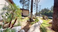This ground-floor residential unit is part of a multi-building apartment complex set within a lush, landscaped environment characteristic of Southern California. The exterior of the complex is finished in warm earth-toned stucco, complemented by wood-accented balconies and open stairwells that connect the upper floors of the surrounding buildings. The unit itself is accessed via a private entry door framed by stucco walls, flanked by windows fitted with vertical blinds and a wall-mounted air conditioning unit, providing climate control suited to the warm regional climate. The grounds of the complex are generously landscaped, featuring tall palm trees, mature pines, tropical shrubs, and decorative boulders that lend a naturalistic character to the outdoor environment. A curving concrete walkway winds through the manicured lawn, creating an inviting approach to the buildings. The interior courtyard between structures is planted with dense tropical foliage, including arching palm varieties, creating a secluded and shaded outdoor corridor. A site map sign near the entrance identifies the complex layout, and wrought iron security gates with ornamental detailing provide controlled access points throughout the property. The overall ambiance reflects a well-established residential community with mature landscaping and a relaxed, sun-drenched atmosphere.