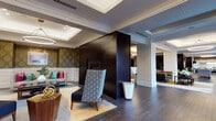 This sophisticated single-floor space showcases an exceptional blend of classical architectural elements and modern interior design. The entrance greets visitors with bold black double doors framed by ornate gold damask wallpaper, crisp white wainscoting, and a dramatic circular pendant light suspended from a coffered ceiling — establishing a refined and welcoming tone throughout.

The expansive lounge area is anchored by a striking dark-paneled fireplace column that serves as a functional room divider, featuring a wall-mounted television on one side. Seating is generously arranged with plush sofas and swivel accent chairs in muted greens, grays, and yellows, all resting on richly patterned area rugs atop dark hardwood floors. Layered, dimmable ring pendant lights contribute to the warm, inviting ambiance.

A formal dining area accommodates a large dark wood table surrounded by upholstered chairs, complemented by a vibrant red bench, an oversized abstract artwork, and a row of large windows that allow abundant natural light and offer views of the outdoors. Multiple pendant lights hang gracefully overhead.

The kitchen is sleek and modern, featuring dark cabinetry, stainless steel appliances, under-cabinet lighting, and a generous island topped with dramatic veined marble — providing both functionality and visual impact. Glass partition walls integrate the kitchen seamlessly into the broader space.