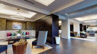 This sophisticated single-floor space showcases an exceptional blend of classical architectural elements and modern interior design. The entrance greets visitors with bold black double doors framed by ornate gold damask wallpaper, crisp white wainscoting, and a dramatic circular pendant light suspended from a coffered ceiling — establishing a refined and welcoming tone throughout.

The expansive lounge area is anchored by a striking dark-paneled fireplace column that serves as a functional room divider, featuring a wall-mounted television on one side. Seating is generously arranged with plush sofas and swivel accent chairs in muted greens, grays, and yellows, all resting on richly patterned area rugs atop dark hardwood floors. Layered, dimmable ring pendant lights contribute to the warm, inviting ambiance.

A formal dining area accommodates a large dark wood table surrounded by upholstered chairs, complemented by a vibrant red bench, an oversized abstract artwork, and a row of large windows that allow abundant natural light and offer views of the outdoors. Multiple pendant lights hang gracefully overhead.

The kitchen is sleek and modern, featuring dark cabinetry, stainless steel appliances, under-cabinet lighting, and a generous island topped with dramatic veined marble — providing both functionality and visual impact. Glass partition walls integrate the kitchen seamlessly into the broader space.