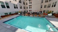 This outdoor amenity space is designed as a central courtyard enclosed by the crisp white facades of a modern multi-story building. The layout is thoughtfully organized into several distinct zones, each serving a different leisure function while maintaining a cohesive, contemporary aesthetic.

At the heart of the space is a generously sized rectangular swimming pool with clear turquoise water, finished with tiled accents and surrounded by a clean concrete deck. Adjacent to the pool, a sun deck features rows of cushioned chaise lounges positioned beneath open sky, with string lights overhead and glimpses of palm trees visible above the roofline, lending a distinctly urban resort ambiance.

A dedicated lounge area centers around a sleek rectangular fire pit table with glass bead fill, flanked by wicker seating with colorful accent cushions and rounded ottomans in warm orange. This seating zone connects to an elevated dining terrace via a wood-and-glass staircase, adding architectural dimension to the courtyard.

The outdoor kitchen and dining area is well-equipped with a built-in gas grill, prep sink, and rustic wood cabinetry, accompanied by multiple dining tables shaded by bold red-and-white striped umbrellas. Potted plants and sculptural orange planters add pops of color throughout, completing the inviting, resort-style atmosphere.