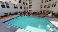 This outdoor amenity space is designed as a central courtyard enclosed by the crisp white facades of a modern multi-story building. The layout is thoughtfully organized into several distinct zones, each serving a different leisure function while maintaining a cohesive, contemporary aesthetic.

At the heart of the space is a generously sized rectangular swimming pool with clear turquoise water, finished with tiled accents and surrounded by a clean concrete deck. Adjacent to the pool, a sun deck features rows of cushioned chaise lounges positioned beneath open sky, with string lights overhead and glimpses of palm trees visible above the roofline, lending a distinctly urban resort ambiance.

A dedicated lounge area centers around a sleek rectangular fire pit table with glass bead fill, flanked by wicker seating with colorful accent cushions and rounded ottomans in warm orange. This seating zone connects to an elevated dining terrace via a wood-and-glass staircase, adding architectural dimension to the courtyard.

The outdoor kitchen and dining area is well-equipped with a built-in gas grill, prep sink, and rustic wood cabinetry, accompanied by multiple dining tables shaded by bold red-and-white striped umbrellas. Potted plants and sculptural orange planters add pops of color throughout, completing the inviting, resort-style atmosphere.