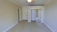 This single-floor space presents a clean, functional layout highlighted by several classic architectural details. The main room is finished with neutral-toned carpet and warm beige walls, creating a calm, understated ambiance throughout. Crown molding runs along the ceiling perimeter, adding a traditional, refined character to the interior. A ceiling fan with a light fixture is centrally mounted, providing both air circulation and ambient lighting.

A prominent feature of the room is the built-in closet, fitted with mirrored sliding doors that visually expand the sense of space. Inside, built-in shelving offers organized storage. Adjacent to the closet, a solid entry door with gold-toned hardware provides access to the space, while a secondary doorway leads to an additional room or hallway, suggesting a practical, connected floor plan.

The white trim work throughout — including baseboards, door casings, and molding — contrasts cleanly against the neutral walls, reinforcing the space's traditional aesthetic. Overall, the interior conveys a straightforward, well-maintained character suited to comfortable, everyday living.