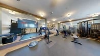 The fitness room at 619 North in Ho-Ho-Kus, New Jersey, is a thoughtfully designed and comprehensively equipped exercise facility spread across a single floor. The space is organized into distinct functional zones, seamlessly flowing from one area to the next.

The cardio zone lines one side of the room, featuring a row of treadmills and elliptical trainers equipped with individual display screens. The herringbone-patterned hardwood-style flooring in this section lends a refined aesthetic, complemented by bold black-and-white abstract artwork adorning the walls. Flush-mount globe lighting illuminates the space with a clean, modern glow.

Transitioning into the strength training area, the flooring shifts to durable rubber matting suited for heavier use. This section houses a multi-use cable machine, adjustable weight benches, a full rack of dumbbells, and a branded functional training rig stocked with kettlebells, medicine balls, battle ropes, and a plyo box. Natural light filters in through windows, brightening the strength zone alongside the overhead lighting.

A dedicated functional and cycling area features spin bikes, a BOSU trainer, and a large flat-screen television mounted against mirrored walls that visually expand the space. The mirrors also reflect the well-organized layout, reinforcing the room's spacious and open feel. Overall, the fitness room combines practical, high-quality equipment with polished, modern design elements.