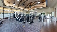 This fitness center in Royal Palm Beach, Florida, offers a generously sized, single-floor facility designed to support a full range of workout routines. The main gym area is architecturally distinguished by soaring vaulted ceilings with exposed wooden beam trusses and stylish pendant lighting, creating an open and energizing atmosphere. Large, multi-pane windows line the walls, flooding the space with abundant natural light and providing views of the surrounding greenery. The floor is finished with durable, dark wood-tone and carpet tile surfaces suited for high-traffic use. The main floor is outfitted with a comprehensive selection of Matrix and Matrix-compatible cardio machines, including a row of treadmills, ellipticals, and stationary bikes, alongside a full lineup of cable and resistance strength machines. A dedicated studio space features warm hardwood flooring, soaring ceilings with clerestory windows, full-length mirrored walls, and glass partitions. It is equipped with spin bikes, medicine balls, stability balls, and a dumbbell rack, making it ideal for group classes or focused individual training. A wall-mounted flat-screen TV enhances the studio experience. Throughout the facility, the design balances modern finishes with a warm, inviting aesthetic, making it a functional and visually appealing environment for fitness.