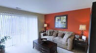 This two-floor residential space is designed with comfort and style in mind, offering a well-appointed layout across two levels. The living area immediately draws attention with its striking terracotta-toned accent wall, which adds warmth and character to the room. Soft, plush carpeting underfoot enhances the cozy atmosphere, while a large sliding glass door fitted with vertical blinds allows for generous natural light and provides direct access to the outdoors. The living space is generously proportioned, easily accommodating a full-sized sofa suite and a substantial dark wood coffee table with built-in storage drawers, contributing both function and elegance to the room. Neutral tones on the surrounding walls complement the bold accent, creating a balanced and welcoming ambiance throughout. The overall design aesthetic blends contemporary touches with classic comfort, making the space feel both livable and visually engaging. With its two-floor layout, the home offers a natural separation between living and private spaces, providing a practical and comfortable flow for everyday living.
