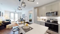 This single-floor residence showcases a cohesive modern design with a neutral palette of whites and soft grays accented by warm wood tones. The open-concept kitchen is a central highlight, featuring gray shaker-style cabinetry extending to the ceiling, stainless steel appliances, white quartz countertops, a deep undermount sink, and a spacious island with seating. Geometric black pendant lights hang above the island, adding a contemporary industrial touch. The kitchen flows seamlessly into the living area, which benefits from large floor-to-ceiling windows and a sliding glass door that invites abundant natural light.

The primary bedroom offers a comfortable retreat with plush carpeting, a ceiling fan, and direct access to a generously sized walk-in closet outfitted with multiple hanging rods and built-in shelving. A second bedroom provides additional flexibility, also finished with carpet and ample space. The bathroom is well-appointed with gray vanity cabinetry, a white quartz countertop, a large mirror, tile flooring, and a full tub with subway tile surround. A wide hallway connects the private areas of the residence, finished with large-format tile and additional storage access, reinforcing the space's functional and polished character.
