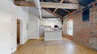 Located at 237-243 Furnace St in Akron, OH, this one-bedroom, one-bathroom loft masterfully blends industrial heritage with contemporary living. The open-concept main area showcases soaring ceilings with exposed black-painted structural rafters, original heavy timber beams, and visible ductwork — all hallmarks of a thoughtfully converted historic building. A striking exposed brick wall runs throughout the space, adding warmth and texture.

The kitchen is well-appointed with stainless steel appliances — including a refrigerator, electric range, microwave, and dishwasher — paired with white upper cabinetry, warm wood lower cabinets, a tile backsplash, and light quartz countertops. A central island provides additional prep and dining space, while pendant lighting adds a modern industrial touch. Light hardwood flooring grounds the main living and kitchen areas.

The bedroom offers a comfortable footprint with plush carpeting, a double-door closet, and the same dramatic exposed brick and original timber beams overhead, with a large arched window allowing natural light to fill the room. The bathroom is clean and modern, featuring a tiled soaking tub and shower combo, wood-toned vanity, quartz countertop, and hardwood flooring — providing a calm, refined retreat within this characterful space.